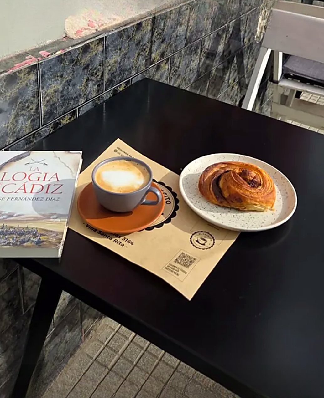 Un libro, un café con leche y una factura sobre una mesa negra, junto a la ventana