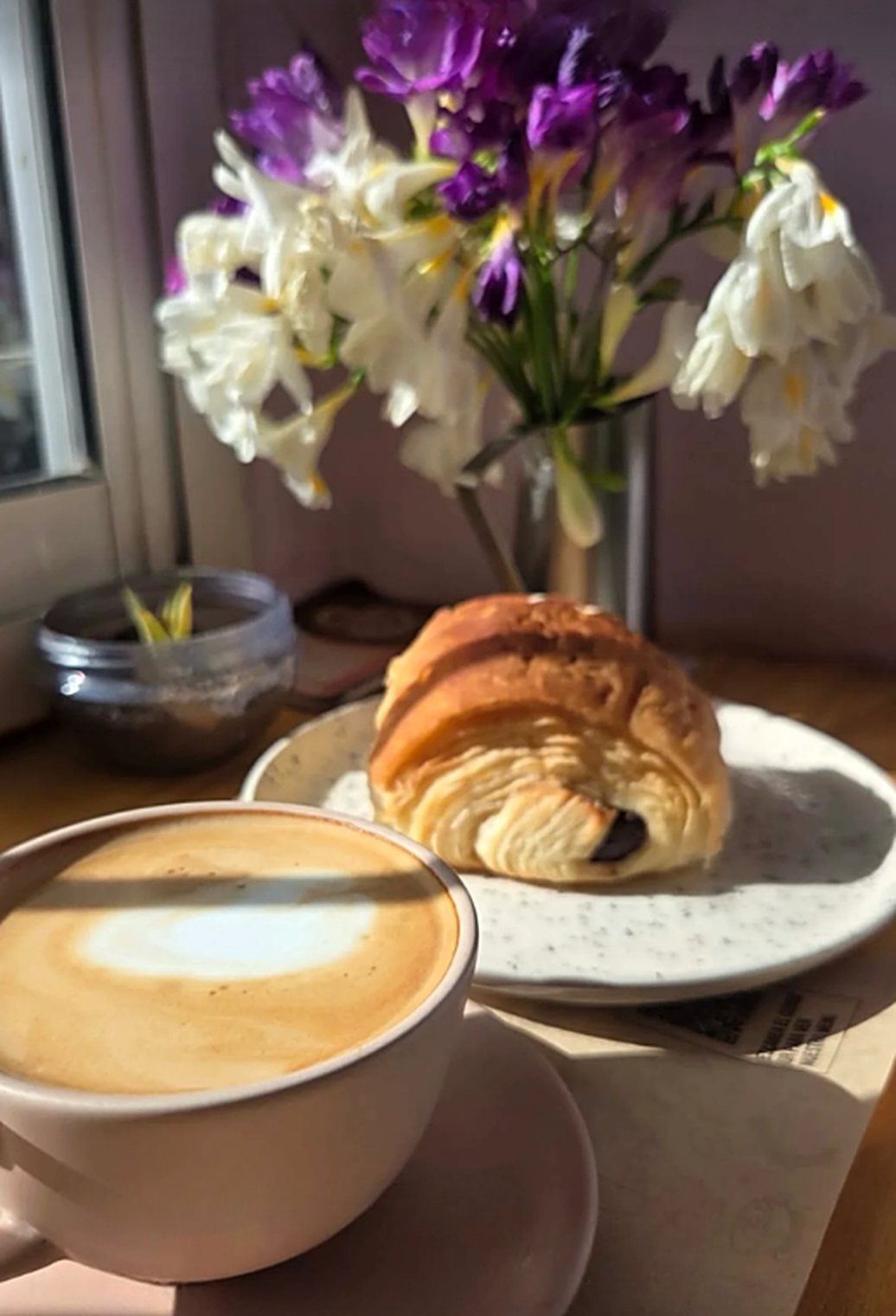 Café con leche y una factura de chocolate al sol, junto a un florero con fresias
